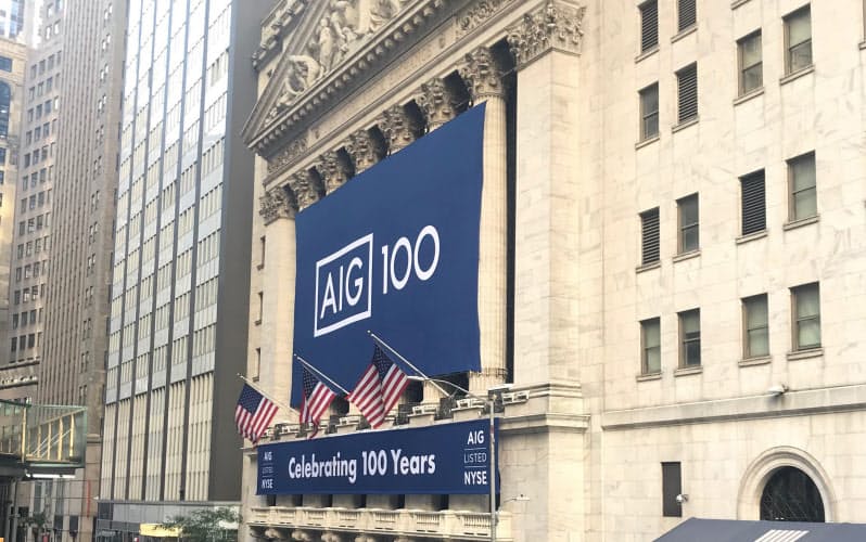 AIGもダイベストメントの波にさらされる(ニューヨーク証券取引所に掲げられた創立100周年を祝うバナー)=ロイター AIGもダイベストメントの波にさらされる(ニューヨーク証券取引所に掲げられた創立100周年を祝うバナー)=ロイター