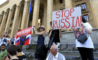 トビリシのジョージア議会前で「ロシアを止めろ」と書かれたプラカードを掲げるデモ参加者ら(6月20日)=AP トビリシのジョージア議会前で「ロシアを止めろ」と書かれたプラカードを掲げるデモ参加者ら(6月20日)=AP