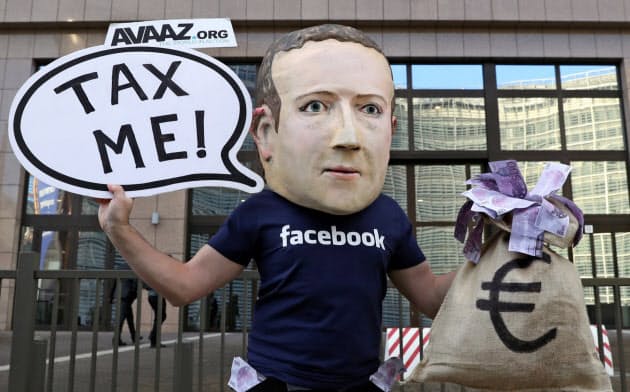 EU本部前でフェイスブックのザッカーバーグCEOにふんしてデジタル課税を訴える人物=ロイター EU本部前でフェイスブックのザッカーバーグCEOにふんしてデジタル課税を訴える人物=ロイター