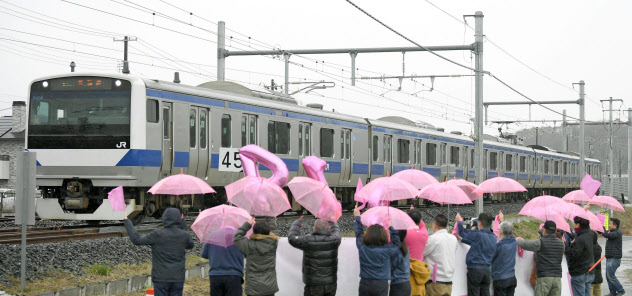 JR常磐線が全線運行再開し、富岡―夜ノ森間を走行する列車。町民らが桜をイメージしたピンクの傘などで歓迎した(14日、福島県富岡町)=共同 JR常磐線が全線運行再開し、富岡―夜ノ森間を走行する列車。町民らが桜をイメージしたピンクの傘などで歓迎した(14日、福島県富岡町)=共同