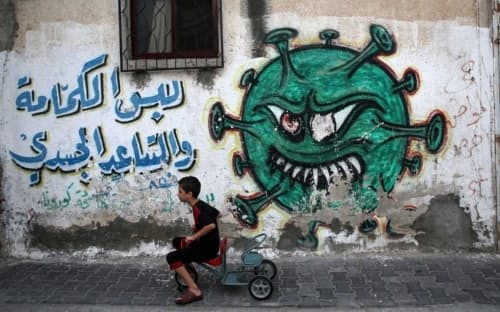 2022年8月29日、パレスチナのガザ市にあるパレスチナ難民のためのアルシャティキャンプで、ウイルスを描いた壁画と新型コロナウイルスの予防策を促すメッセージの前を三輪車で通り過ぎる少年。（PHOTOGRAPH BY MAJDI FATH, NURPHOTO/GETTY IMAGES）