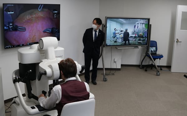 東京の熟練の医師（左手前）が、愛知の若手医師（右奥画面）のロボット手術を指導する