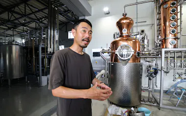 蒸留器を前に事業展開を説明する熊本ワインファームの西村氏(熊本県山鹿市、7月)