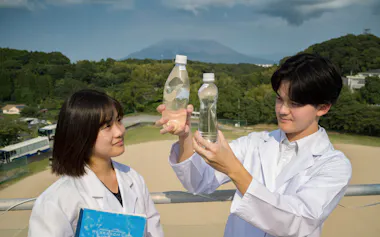 桜島(後方)で採取した温泉水を手にする池田学園池田高校の茶屋道玲さん㊨と川路眞愛さん